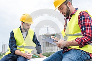 two engineers in safety vests and hardhats working with blueprints and clipboard