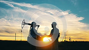 Two engineers shaking hands in front of oil pumpjack at sunset background