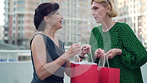 Two envious adult women bragging purchases in shopping bags talking standing on city street. Portrait of Caucasian