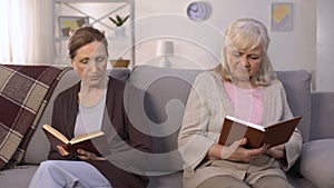 Two envious old women reading books sitting on hall of nursing home, rivalry