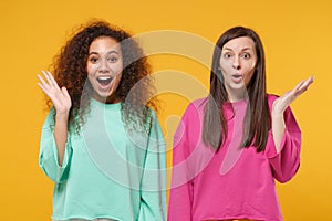 Two excited amazed women friends european and african american girls in pink green clothes posing isolated on yellow
