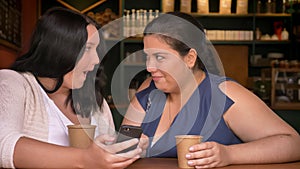Two excited overweighted caucasian girls flancing at phone holding cuos of coffee in cafe