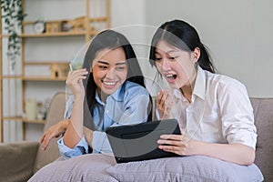 Two excited young women using a digital tablet together while relaxing on couch in the living room at home.