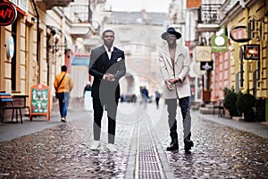 Two fashion black men walking on street. Fashionable portrait of african american male models. Wear suit, coat and hat photo