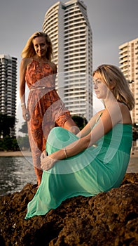 Two fashion models posing in red and turquoise dresses in the rays of the setting sun on tropical beach on a background