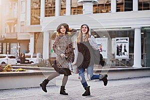 Two fashionable joyful smiling girls jumping over city background. Stylish look, travelling together, wearing modern