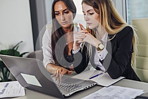 Two female accountants working together on financial report using laptop sitting at desk in account department