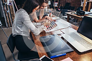 Two female architects working together using color swatches sitting at desk with laptop, graphic tablet in design studio