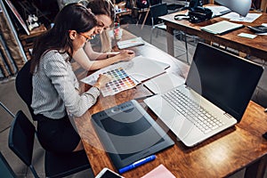 Two female architects working together using color swatches sitting at desk with laptop, graphic tablet in design studio