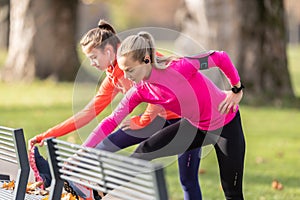 Two female athletes are warming up in the autumn park, using a bench to warm up before running