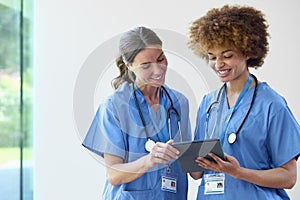 Two Female Doctors Wearing Scrubs With Digital Tablet Discussing Patient Notes In Hospital
