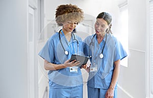 Two Female Doctors Wearing Scrubs With Digital Tablet Discussing Patient Notes In Hospital