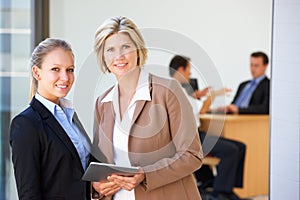 Two Female Executives Using Tablet Computer With Office Meeting In Background