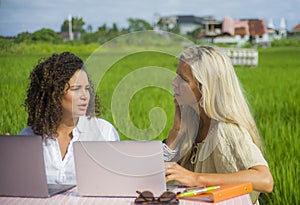 Two female friends caucasian and afro working together outdoors at cool internet cafe with laptop computer as digital nomad trave