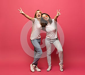 Two female friends hugging and having fun together, showing peace gesture while looking at camera, isolated over pink