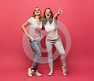 Two female friends hugging and having fun together, showing peace gesture while looking at camera, isolated over pink