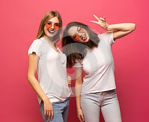 Two female friends hugging and having fun together, showing peace gesture while looking at camera, isolated over pink