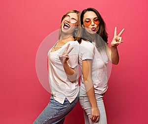 Two female friends hugging and having fun together, showing peace gesture while looking at camera, isolated over pink