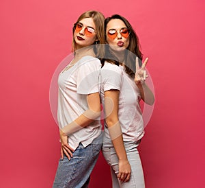 Two female friends hugging and having fun together, showing peace gesture while looking at camera, isolated over pink