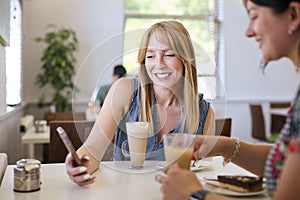 Two Female Friends Meeting In Coffee Shop Looking At Mobile Phone Together