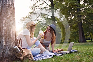 Two female friends sitting on the grass, talking and using laptop