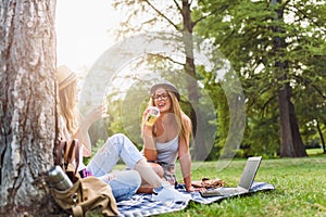 Two female friends sitting on the grass, talking and using laptop