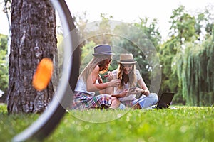 Two female friends sitting on the grass, talking and using laptop