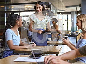 Two Female Friends At Table Meeting In Busy Coffee Shop Talking To Waitress