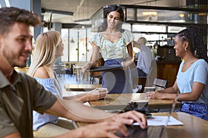Two Female Friends At Table Meeting In Busy Coffee Shop Talking To Waitress