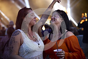 Two Female Friends Wearing Glitter Dancing At Outdoor Summer Music Festival Holding Drinks At Night