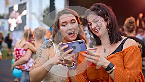 Two Female Friends Wearing Glitter Looking At Mobile Phone At Summer Music Festival Holding Drinks