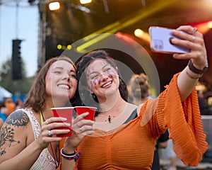 Two Female Friends Wearing Glitter Posing For Selfie At Summer Music Festival Holding Drinks