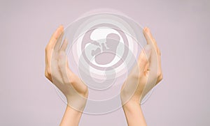 Two female hands hold an icon of a human embryo. Close-up, pink isolated background. The concept of the origin of the world