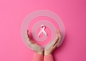 Two female hands hold a pink silk ribbon in the form of a loop on a pink background. Symbol of the fight against breast cancer