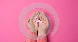 Two female hands hold a pink silk ribbon in the form of a loop on a pink background. Symbol of the fight against breast cancer