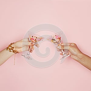 Two female hands holding wine glasses with colorful holiday confetti on pink background. Celebration, party and holidays
