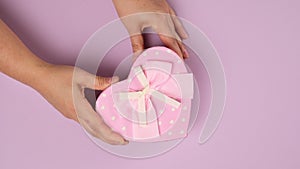 Two female hands put on a lilac background a pink cardboard gift box in a heart shape, top view