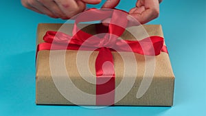 Two female hands tie a red bow on a wrapped box, blue background, concept of congratulations on the holiday