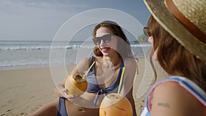 Two female happy friends in bikini tanning, chilling with coconut cocktails at sandy beach. Attractive smiling girls