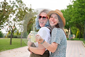 Two female happy friends drinking refreshing drinks, laughing, hugging and looking to the camera in a park