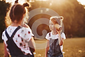 Two female kids stands in the field and talking by using string can phone