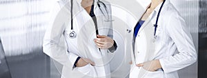 Two female physicians are discussing medical therapy, while standing at the table in a clinic office. Doctors use pc