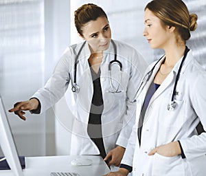 Two female physicians are discussing medical therapy, while standing at the table in a clinic office. Doctors use pc