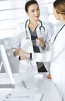 Two female physicians are discussing medical therapy, while standing at the table in a clinic office. Doctors use pc