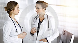 Two female physicians are discussing medical therapy, while standing at the table in a clinic office. Doctors use pc