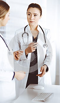 Two female physicians are discussing medical therapy, while standing at the table in a clinic office. Doctors use pc