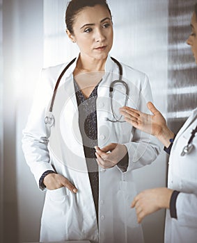 Two female physicians are discussing medical therapy, while standing at the table in a sunny clinic office. Doctors at