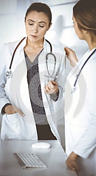 Two female physicians are discussing medical therapy, while standing at the table in a sunny clinic office. Doctors at