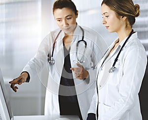 Two female physicians are discussing medical therapy, while standing at the table in a sunny clinic office. Doctors use