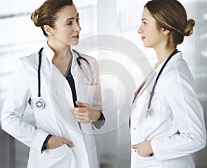 Two female physicians are discussing medical therapy, while standing at the table in a sunny clinic office. Doctors use